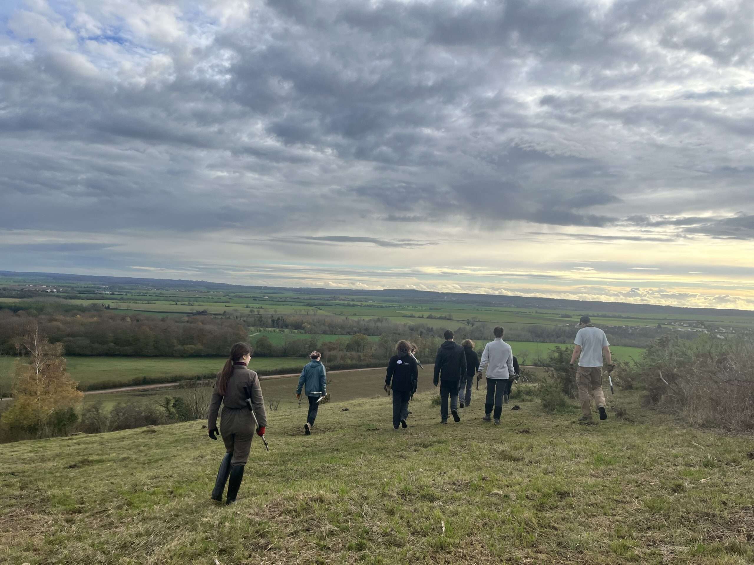 Personnes marchant sur un coteau herbeux au Mesnil-Soleil, équipées d’outils pour un chantier nature, avec vue dégagée sur la vallée et ciel nuageux en arrière-plan.