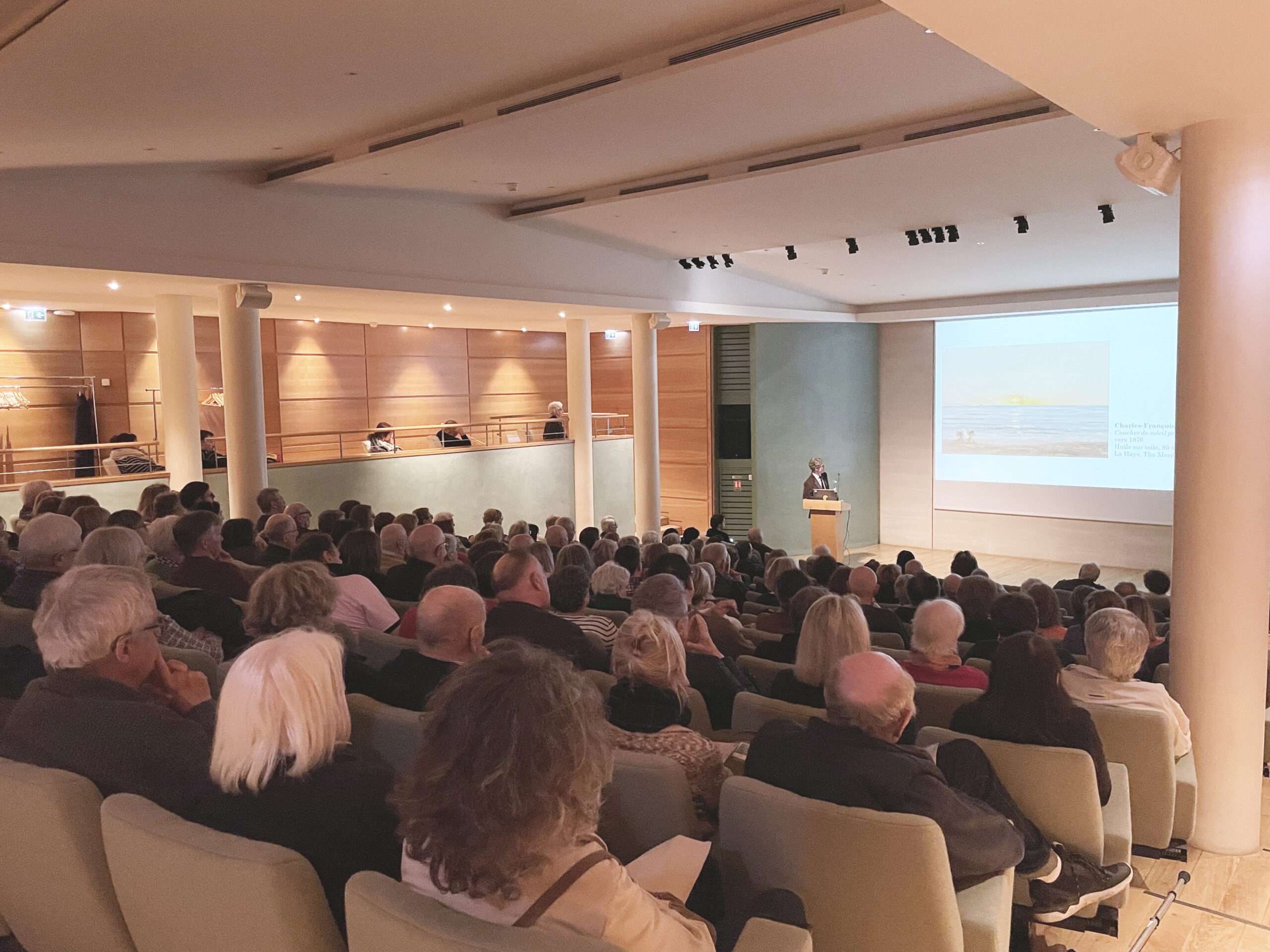 Vue de dos d'un public attentif assis dans l'auditorium du musée des impressionnismes Giverny. Un conférencier se tient derrière un pupitre face à l'assistance, tandis qu'un grand écran projette la peinture d'un paysage marin, illustrant l'analyse des œuvres de Monet et de ses contemporains.